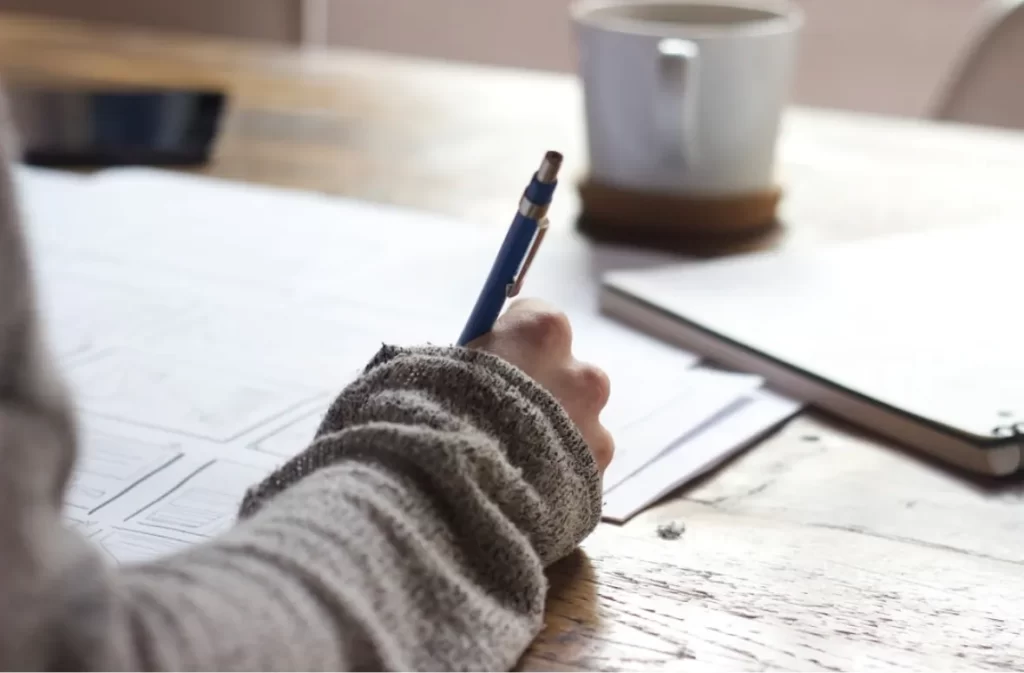 Person writing essay with blue pen on desk.
