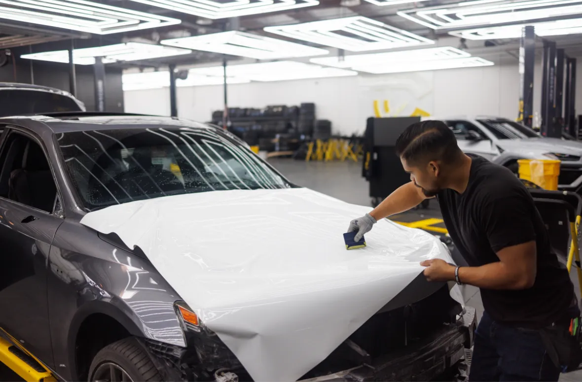 Worker smoothing white wrap on a car hood in a garage.