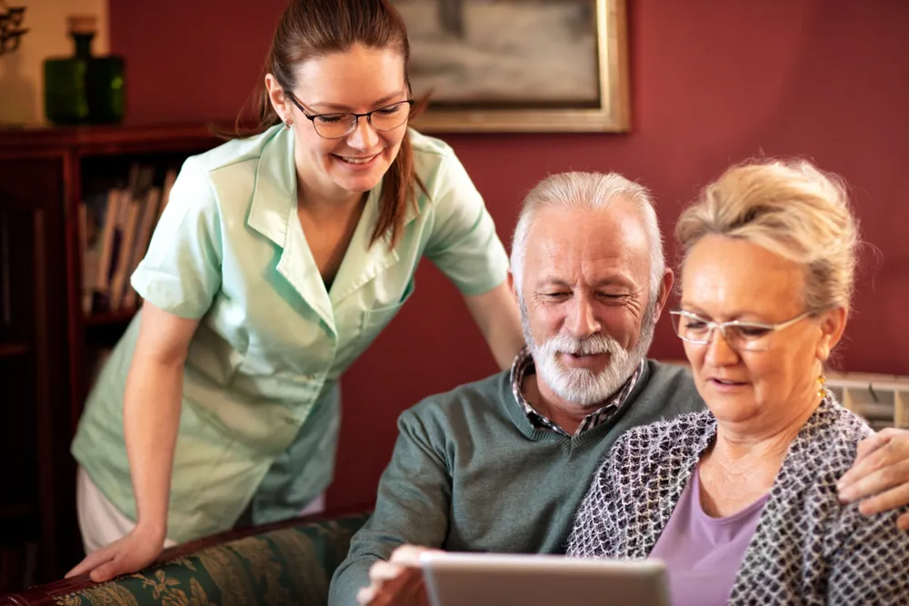 A caregiver helps an elderly couple with technology at home, looking at a tablet together in a cozy living room.